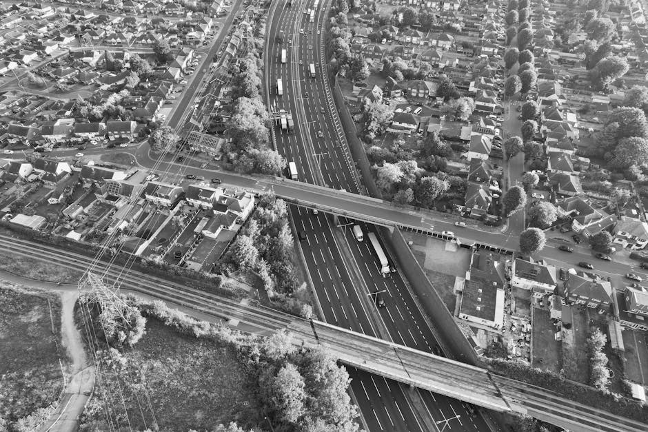 An aerial black-and-white photograph showing a section of an urban street with multiple lanes of traffic and a bridge crossing over a railway line. The scene includes a network of roads, some with trucks and cars moving along them, and trees lining the streets and surrounding residential houses. The houses are densely packed, with gardens and driveways visible, and the area features a mixture of rooftops, some with chimneys, and streets with asphalt road surfaces marked with lane lines. The bridge structure appears to be a key part of the street-level logistics involved in home relocation or furniture transport, with a few visible vehicles possibly involved in packing and moving processes. Man With a Van Brent Cross may utilize this route for efficient removals near Brent Cross, with the highway and street infrastructure supporting loading and unloading activities for house removals, and the railway line indicating local transport connections. The lighting is even, highlighting the detailed layout of the neighborhood and transport routes.