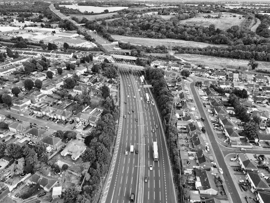 A black-and-white aerial photograph showing a multi-lane motorway with moving vehicles including cars, trucks, and vans, running through a suburban area with houses, gardens, and trees on either side. The motorway crosses under a bridge that connects two sections of the road and is surrounded by residential streets lined with detached and semi-detached houses, many with visible driveways and small gardens. In the background, fields, trees, and roads extend into the distance under a partly cloudy sky. The scene captures the transportation infrastructure and residential environment typical of a home relocation or furniture transport process, as seen in the context of house removals services offered by Man With a Van Brent Cross.