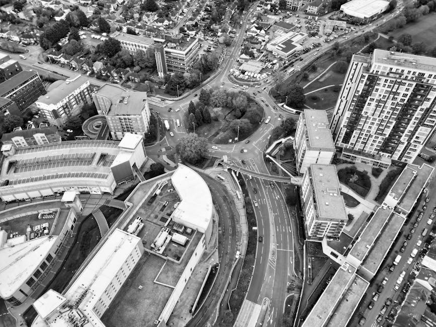 An aerial black-and-white photograph showing a section of an urban street with multiple lanes of traffic and a bridge crossing over a railway line. The scene includes a network of roads, some with trucks and cars moving along them, and trees lining the streets and surrounding residential houses. The houses are densely packed, with gardens and driveways visible, and the area features a mixture of rooftops, some with chimneys, and streets with asphalt road surfaces marked with lane lines. The bridge structure appears to be a key part of the street-level logistics involved in home relocation or furniture transport, with a few visible vehicles possibly involved in packing and moving processes. Man With a Van Brent Cross may utilize this route for efficient removals near Brent Cross, with the highway and street infrastructure supporting loading and unloading activities for house removals, and the railway line indicating local transport connections. The lighting is even, highlighting the detailed layout of the neighborhood and transport routes.