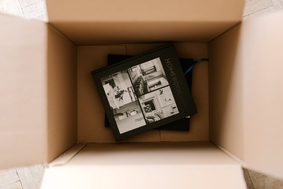 A man wearing a black cap, white polo shirt, and jeans is kneeling on the pavement outside a residential property during daylight, surrounded by several large cardboard moving boxes. He is using a bla