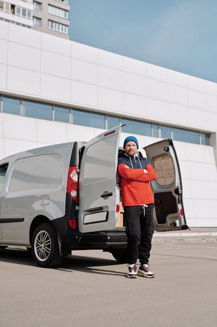 A small red flatbed van parked on the street outside a modern office building with large glass windows and concrete facade. The van's cargo area is loaded with various household items, including wrapp