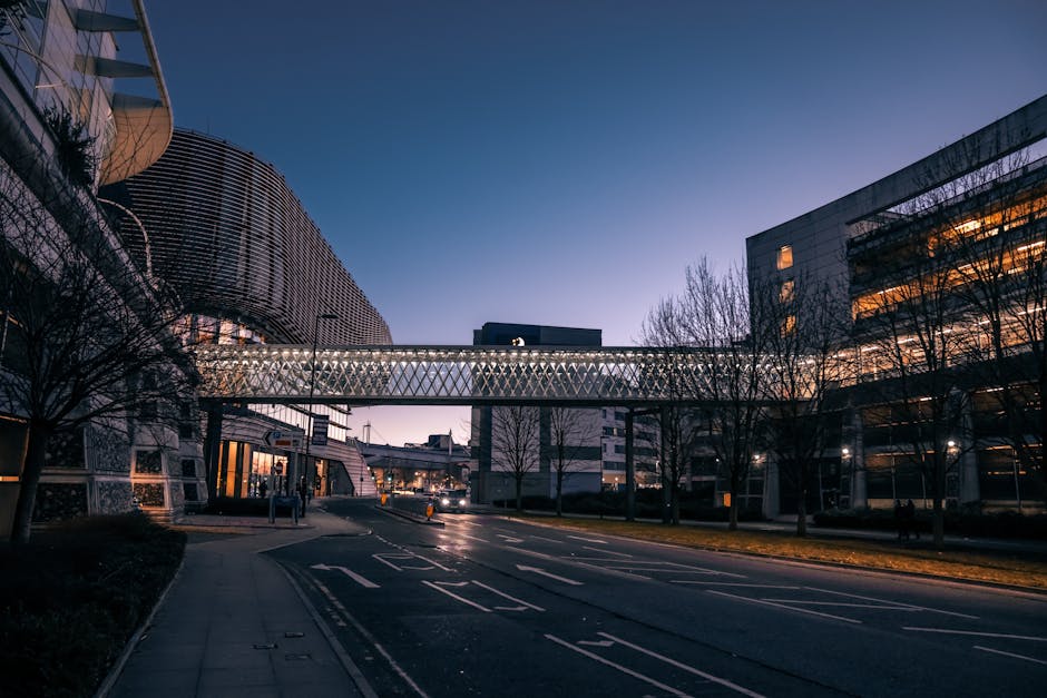 A wide view of a modern urban street at dusk, featuring a pedestrian bridge with a lattice design crossing over the road, connecting two large office buildings with glass facades. The street is lined with leafless trees and illuminated by streetlights, with some building interior lights visible through the windows. The road has multiple lanes and a few parked vehicles in the distance. The scene captures the quiet atmosphere of a city area, which could be part of a home relocation route. Momentarily, no vehicles or moving equipment are visible, but the environment represents an area where furniture transport and packing logistics from a house removal service like Man With a Van Brent Cross may occur in preparation for a house move or office relocation.