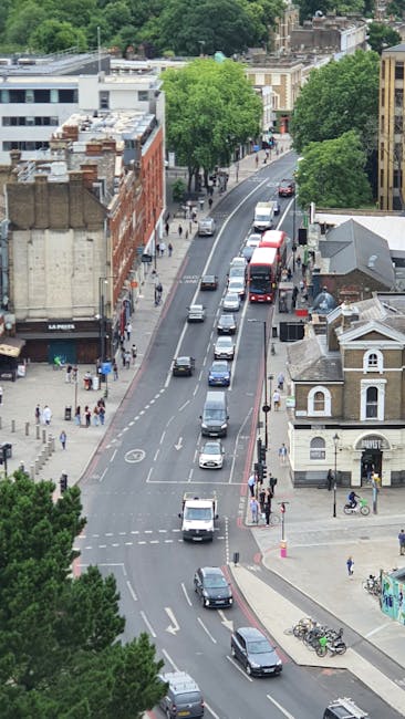 An aerial view of a busy urban street with parked cars lining both sides and moving vehicles in transit, including a red double-decker bus and several smaller cars. The street is flanked by various buildings such as a commercial property with signage and residential houses, some with flat roofs and chimney stacks. Tall, leafy green trees are visible along the sidewalk, providing shade and greenery to the scene. Pedestrians can be seen walking along the pavements, and bicycles are parked near a bicycle rack at the corner. The environment appears well-lit with natural daylight, indicating daytime conditions. This scene illustrates a typical residential and commercial area suitable for home relocation and furniture transport, where careful loading and packing by professional removals services like Man With a Van Brent Cross are essential for smooth moving processes within the Claremont Road, Brent Cross area.