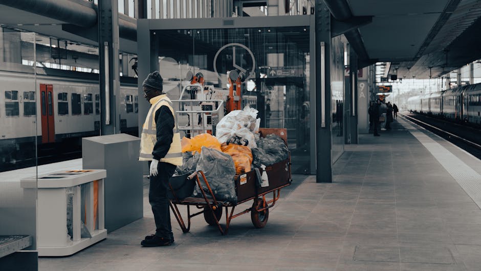A spacious indoor shopping mall with a high, industrial-style ceiling featuring exposed metal trusses and large glass windows allowing natural light to fill the space. The ground floor is occupied by